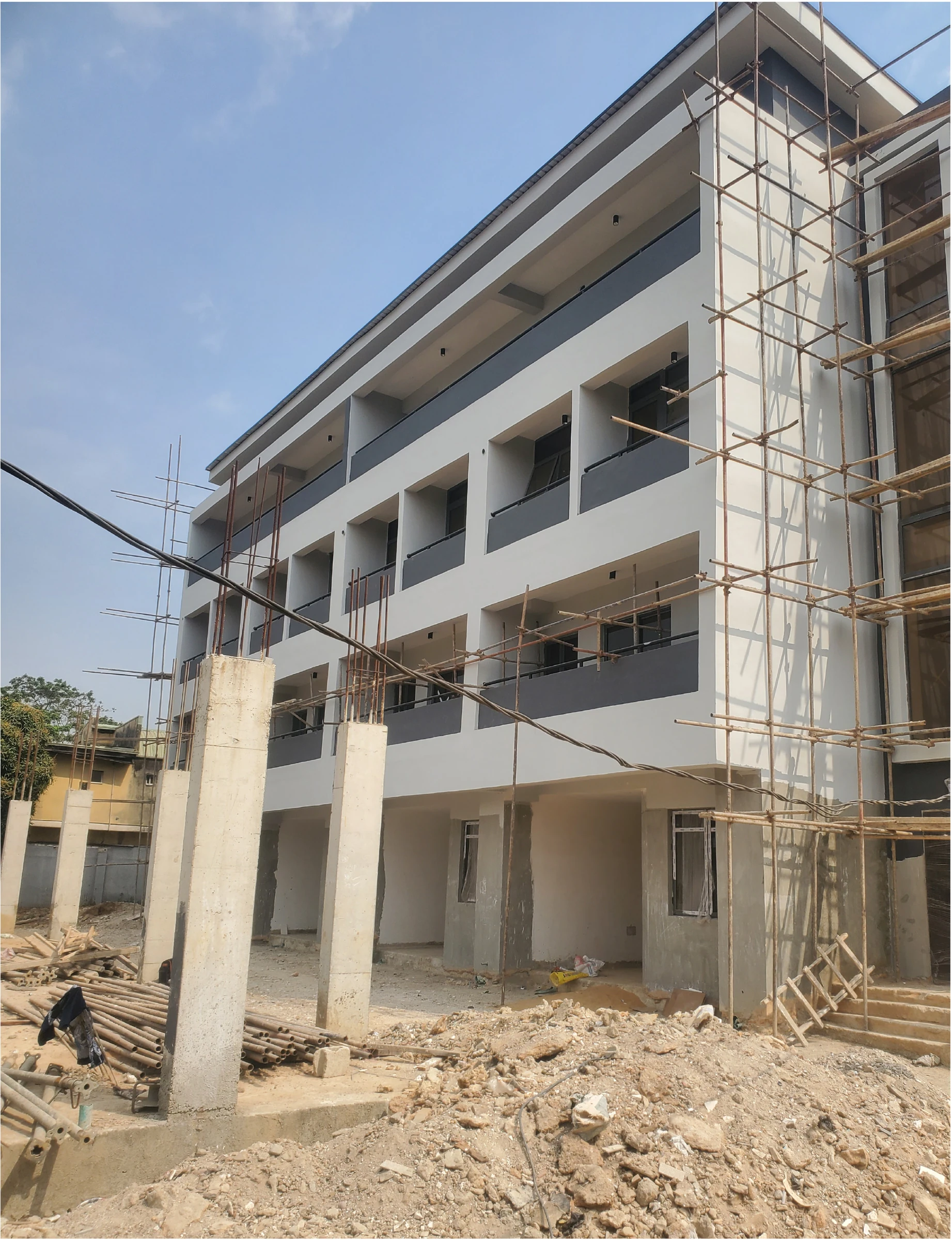Site photograph of the rendered four-storey terrace block with dark balcony bands, scaffolding and concrete columns in the foreground